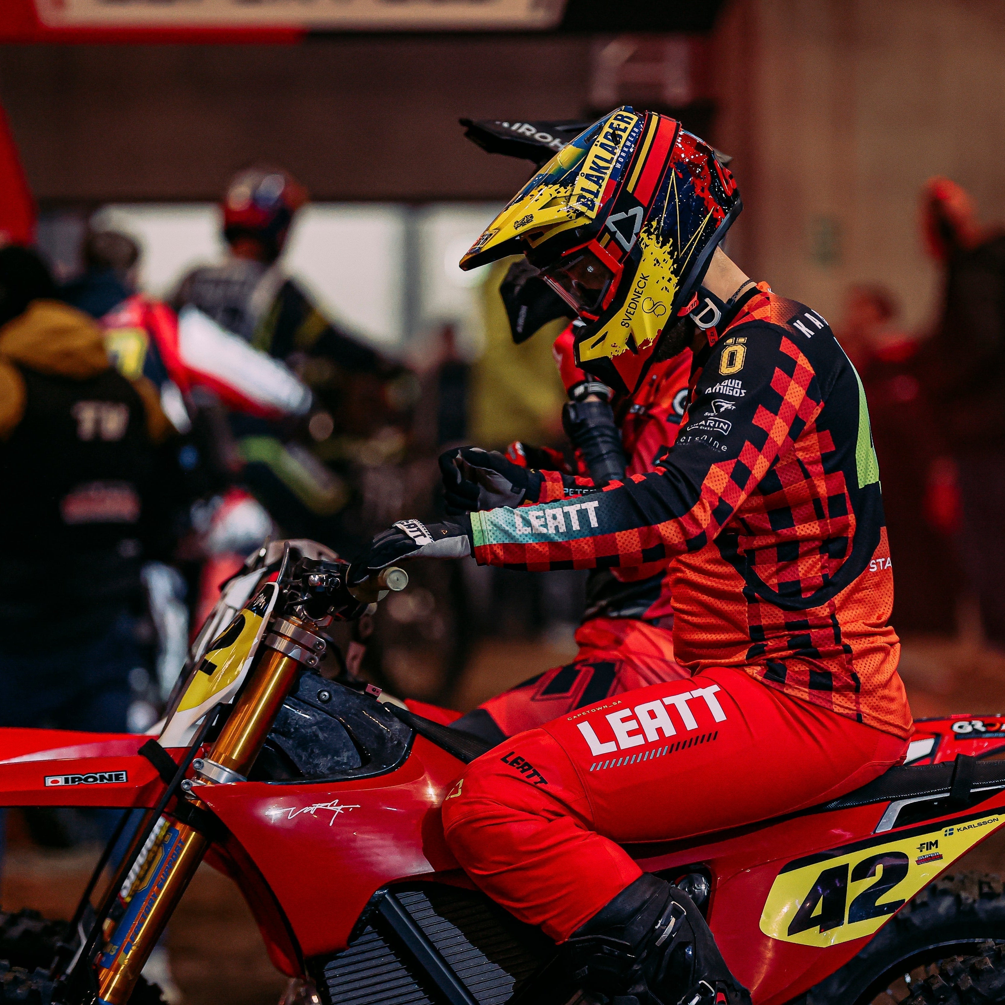 Motorcycle rider in red and black gear with helmet on a dirt bike at an event.